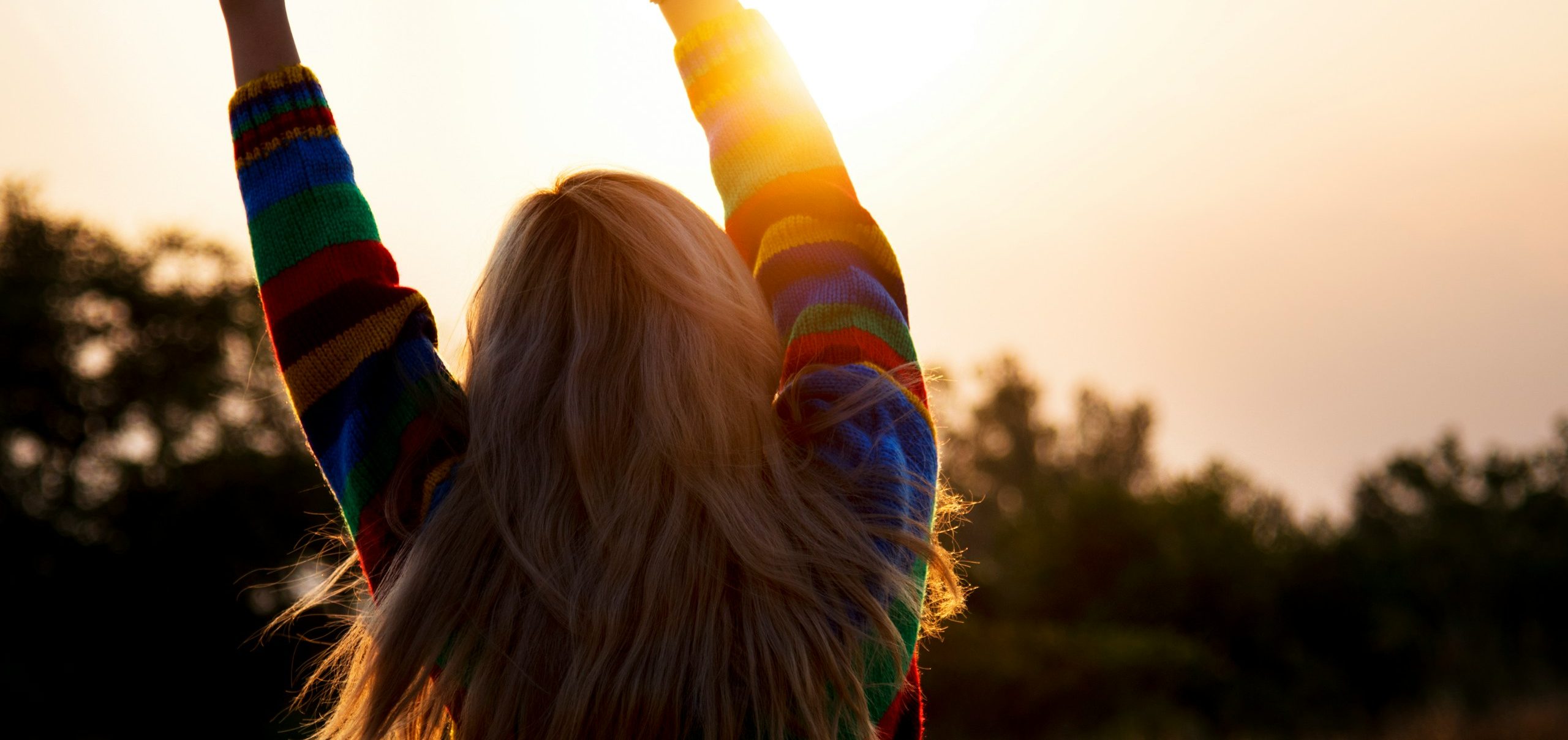 Frau mit buntem Pullover, die die Arme zum Sonnenuntergang hebt.
